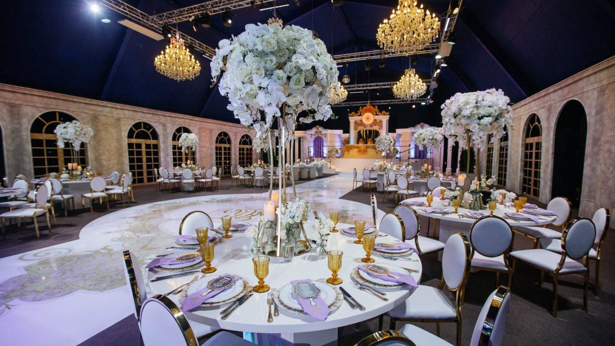 Large white bouquets of roses stand on dinner tables decorated with golden glasses and violet cloth